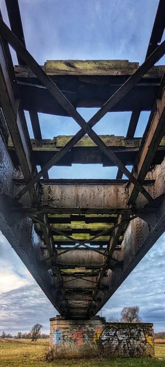 A low-angle shot from directly beneath a decaying railway bridge in Biederitz, Germany, looking up at its dark, rusted metal girders and wooden crossbeams. The intricate, geometric structure of the bridge fills the upper part of the frame, with patches of bright blue sky and white clouds visible through the gaps. The concrete pier supporting the bridge is covered in vibrant graffiti, contrasting with the muted tones of the bridge itself and the grassy, flat landscape beneath it.