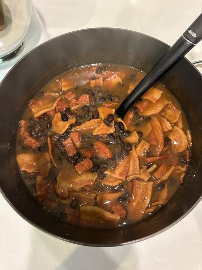 A steaming pot of black beans, chorizo, and sliced bacon in a rich, brown broth. A black ladle rests inside, evoking a warm, hearty meal.