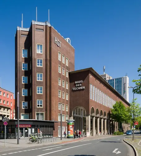 The image features a building complex with a mix of architectural styles. Prominent in the foreground is a structure labeled "HAUS DER TECHNIK," which appears to serve as a center for technology or technical exhibitions. The building has a modern look with large arches and columns. Accompanying this structure is another building that has a red brick facade and is labeled "HOT." The scene is set under a clear blue sky, suggesting a bright day. Overall, it seems to be an urban setting,

Image Credits: Wikimedia / Tuxyso / CC BY-SA 3.0