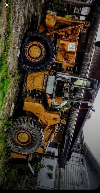 The photograph composed on an Android, shows a large, yellow construction vehicle, likely a front loader or a similar type of heavy machinery, parked on a dirt surface. The vehicle is old and shows significant signs of wear and rust, with its paint chipped and faded in places. The tires are large and rugged, suitable for heavy-duty work, and the vehicle's cabin is partially visible, with the windows and door frame showing signs of age and neglect. The background includes a building with a dark roof and a white wall, suggesting an industrial or rural setting. The sky is overcast, and there is some greenery visible around the vehicle, indicating it is not in a completely barren area. The vehicle is positioned at an angle, with the front facing the right side of the image, and the cabin is on the left side.

 Ovis2-8B

🌱 Energy used: 0.203 Wh