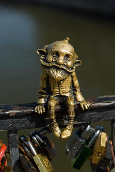 The image captures a whimsical scene featuring a brass sculpture of an elderly man with a large, bushy beard and mustache and prominent glasses. He is dressed in a traditional outfit complete with long-sleeved shirt, checked waistcoat, striped trousers and pointed shoes. The figure is seated comfortably on what appears to be the armrests of a bridge railing.

Under this charming character are multiple padlocks, each differing slightly in color and size, suggesting they may have been placed by various individuals. These locks hang against the backdrop of dark metal railings that frame the scene.

The colors within the photo contrast vividly; the golden hue of brass sculpture stands out starkly against its darker surroundings, drawing attention immediately to it while also serving as a centerpiece for all other objects in view - including those padlocks which add splashes of color and intrigue.