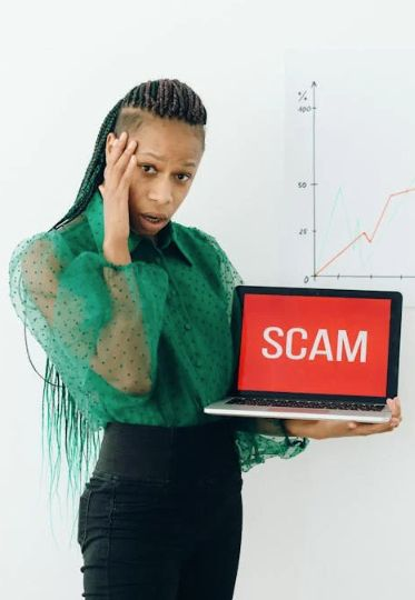 Woman standing with hand to head and other hand holding laptop that says "SCAM". Image by Nataliya Vaitkevich via Pexels