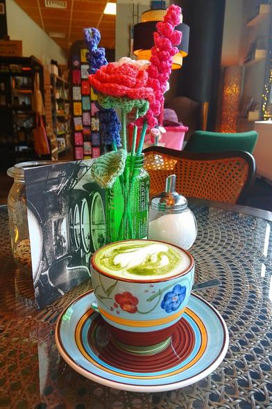 Eine Tasse Matcha Latte in einer blauen Tasse mit roten und blauen Mustern. Diese steht auf einer passenden Untertasse. Tasse und Teller stehen auf einem Glastisch vor einer Flasche mit mit gehäkelten Blumen
