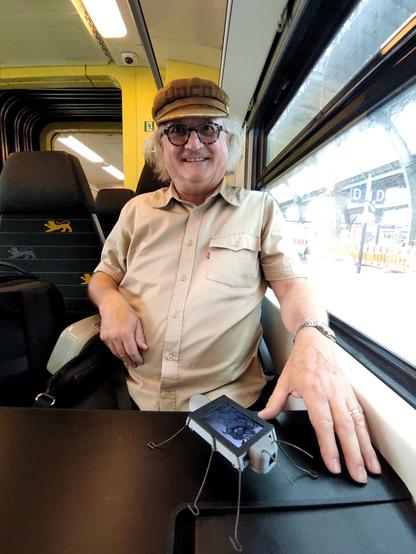 Reinhard is seated at a table in a German regional train. The DigitalBug is standing on the table watching out of the window.