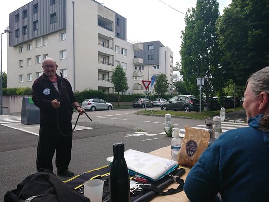 Bénévole de l'association en train de faire le pitre avec un tuyau devant le stand mobile Hérouelibre : table d'outils, boissons et flyers, pied d'atelier avec oriflamme, piste cyclable, rue et immeubles récents à Prestavoine.