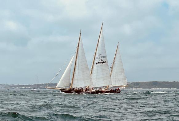 A 19.5 meter sailboat built as a replica of the early 1800s era Bermudian schooners.  She has three masts, with sails set on a windy, overcast day.  The deck is busy with the activities of her racing crew as they set off on a 650 nautical mile race to their home port.