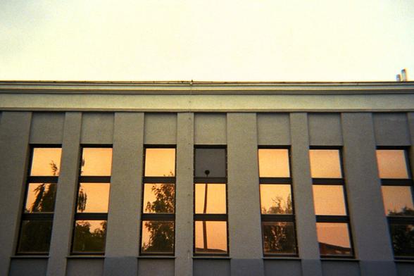 A modern grey building facade with tall rectangular windows reflecting an orange sunset and silhouetted trees. In the central window, the warped reflection of a vertical pole creates a subtle visual tension within the otherwise symmetrical composition