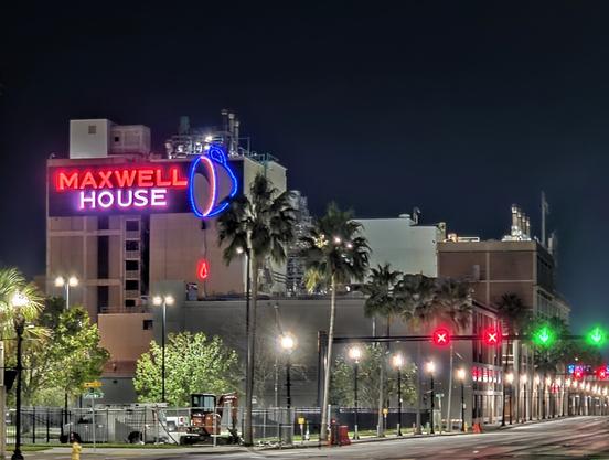 Night view of large coffee factory taking up multiple city blocks, with huge neon sign that reads Maxwell House, with a tipped coffee cup, dripping slowly.