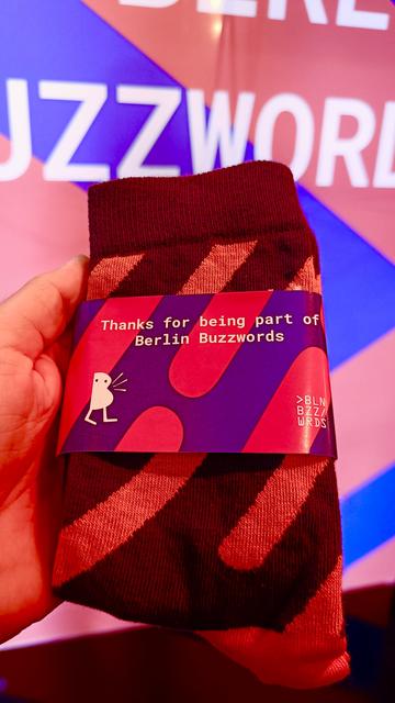 A hand holding a folded pair of purple and orange striped socks. The socks have a label attached that reads "thanks for being a part of Berlin Buzzwords".