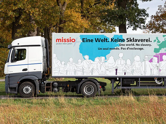 Das Bild zeigt einen weißen Lastwagen mit einem großen Anhänger, der am Straßenrand steht. Im Hintergrund sind Bäume mit herbstlich gefärbtem Laub zu sehen. Auf der Seite des Anhängers ist eine Illustration abgebildet: Silhouetten von Menschen sitzen nebeneinander, dazu ist eine Weltkarte in blauer Farbe zu sehen. Darauf steht auf Deutsch: "Eine Welt. Keine Sklaverei." Darunter steht derselbe Satz auch auf Englisch:
"One world. No slavery." Und auf Französisch: "Un seul monde. Pas d’esclavage." Oben links ist das Logo von „missio“ mit dem Zusatz „glauben.leben.geben.“ zu erkennen. Das Bild ist also eine Werbebotschaft für eine Kampagne, die sich gegen Sklaverei und für Menschenrechte weltweit einsetzt.