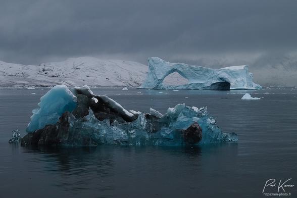 Photo en couleur prise pratiquement au ras de la mer dans un fjord au rivage enneigé, présentant au premier plan un petit grawler aux multiples textures de glace (opaque de couleur blanche à bleu bleue ou transparente, notamment) avec également de la moraine sombre. Ce petit grawler se reflète dans la surface de la mer, pratiquement lisse. Plus loin, on peut voir un iceberg de glace bleue comportant notamment une voûte à gauche ainsi qu'une caverne à droite du côté que l'on peut voir. Encore plus loin, on peut voir des collines enneigées alors que le ciel est complètement bouché par un plafond nuageux relativement bas. Cette lumière relativement parcimonieuse met magnifiquement en valeur les différentes nuances bleues de la glace.