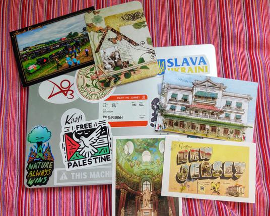 Five postcards from all over the world on top of a bestickerd laptop, lying on a red striped cushion.