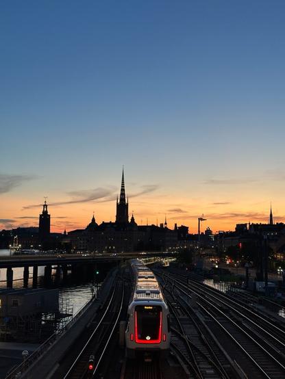 a view over a railway line going towards the silhouette of an old European city, behind which the sun is setting. There is a train passing, its end lit in red, the light of the sunset reflecting off its roof.