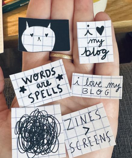 a hand holding tiny handmade stickers. words are spells, zines > screens, i love my blog, and a cat doodle.