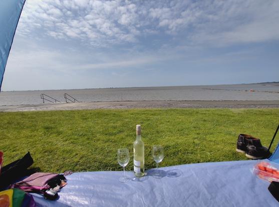 Block aus einer blauen Strandmuschel. Vor ihr erstreckrn sich ca. 5m Wiese, ein Weg und dann die Nordsee. Der Himmel ist blau mit ein paar kleinen Wölkchen. Mittig steht noch eine Flasche alkoholfreier Wein und zwei Gläser in der Strandmuschel.