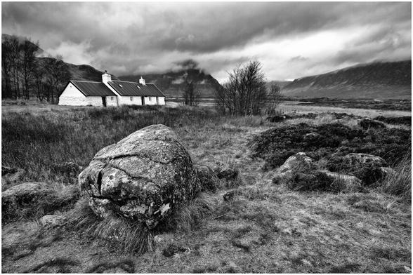 A black and white landscape featuring a rustic cottage surrounded by grassy terrain and a large boulder in the foreground. The sky is overcast with dramatic clouds, and distant mountains are visible in the background.