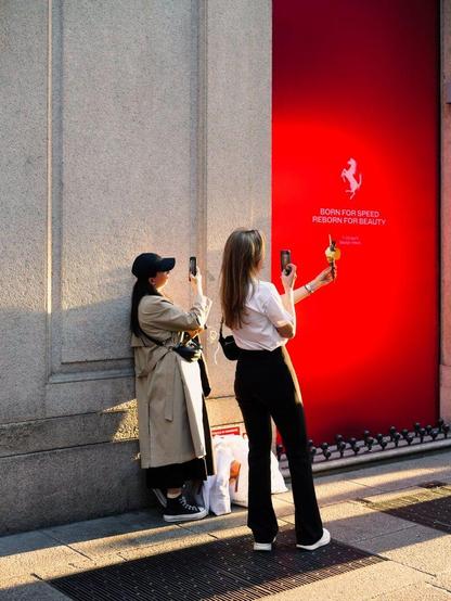 Two young women take photographs of their ice cream against the sunlight near a wall.