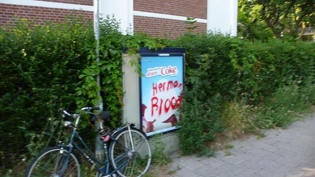 Reclame bord op elektriciteitskastje tussen struiken met een gepakeerde fiets links voor, aangepast met rode letters: herman brood