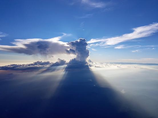 An aerial view shows dramatic clouds with intense sunbeams streaming through them, casting long shadows over a hazy landscape below. The sky is bright blue above the clouds, which are illuminated from behind by the setting or rising sun, creating a striking contrast of light and shadow.