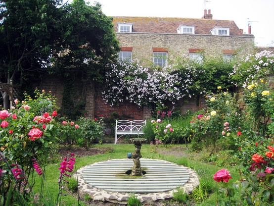 A pretty walled garden with a wrought-iron garden seat, painted white, and a central fountain, with roses and flowering bushes round the sides.