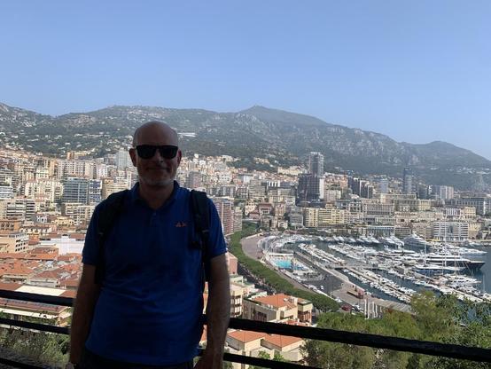 A man wearing sunglasses stands in front of a scenic view of Monaco, featuring a cityscape with various buildings, a marina filled with boats, and mountains in the background under a clear blue sky.