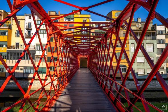 Color photo looking down the enveloping red metal framework of a pedestrian bridge.  Beyond the bridge is a colorful row of riverfront buildings.