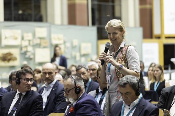 Eine Person steht in einem auditoriumähnlichen Raum und spricht in ein Mikrophon. Die Zuhörer tragen Kopfhörer und schauen zur Bühne, während verschiedene naturkundliche Objekte an der Wand hängen.