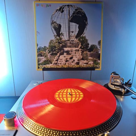 A vinyl record sits on a turntable. Behind the turntable, a vinyl album outer sleeve is displayed. The front cover shows a large globe made of metal sitting on a concrete pedestal that says "Leisure World". There is a small concrete waterfall in front of it