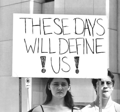 A black and white photo of two people. One holds a handmade sign that says "THESE DAYS WILL DEFINE ! US !" in black letters on a white background. The person holding the sign has dark, shoulder-length hair tucked behind their ears and a solemn look on their face. Another person standing next to them has short dark hair, is wearing oval-shaped wire-framed sunglasses and was caught with their eyes closed and lips pursed as if about to say something. Permission was granted to post this photo. Photo by Jody MacPherson.