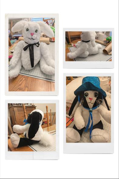 Two handmade stuffed bunnies front view and side view. The top bunny has white fur, green sewn eyes, a sewn pink nose, sewn black mouth, white whiskers and a black ribbon around his neck. The bottom bunny has black and white fur, large green plastic eyes, pink sewn nose, sewn black mouth, white whiskers, blue batik hat with blue ribbon tie with toggle. Both are sitting on a craft work surface with various craft items in the background. Wooden chair tops, table top and wooden antique desk.