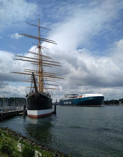 Vorne links das Segelschiff Passat in Travemünde, rechts steuert ein Frachtschiff von der Trave in die Ostsee, an Himmel bergige Wolken.