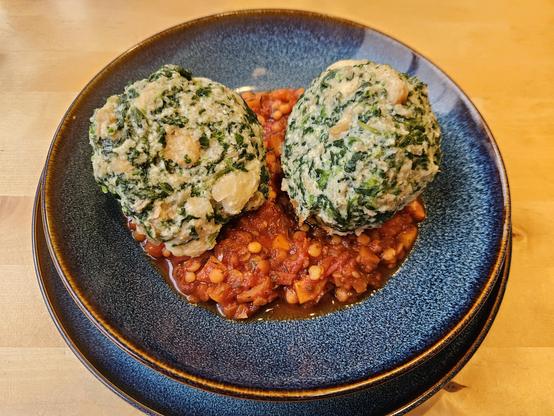 Zwei Spinat-Semmelknödel liegen auf einer Linsen-Paprika-Soße. Serviert auf einem blauen Teller mit braunem Rand.