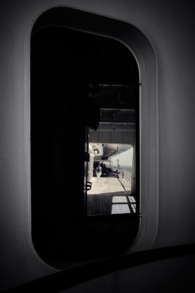 The image shows a view through a series of open doors or windows on a ship. The passage leads to an outdoor deck with railings and a wooden floor. Light creates strong contrasts with shadows, and a person is walking in the distance