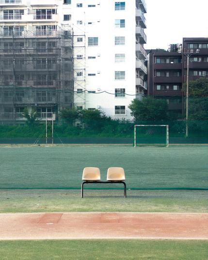 Two cream-colored chairs on a metal frame positioned on a track, facing a fenced soccer field. The field has a goalpost and is partially enclosed with netting. In the background, there are high-rise buildings, some with scaffolding, surrounded by greenery.