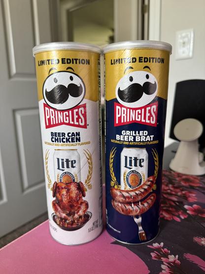 The image features two Pringles cans placed side by side on a table. The can on the left is labeled "Beer Can Chicken" and has a white background with a gold top. It displays an image of a roasted chicken and a can of Miller Lite beer, with the text "Naturally and Artificially Flavored" below the flavor name. The can on the right is labeled "Grilled Beer Brat" and has a dark blue background with a gold top. It features an image of grilled sausages and a can of Miller Lite beer, with the same flavor description text. Both cans are marked as "Limited Edition" at the top. The cans are positioned on a pink tablecloth with a floral pattern, and a white door is visible in the background.