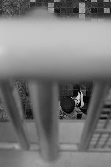 A gentleman in fisherman’s cap, as viewed through railings, passes time.
