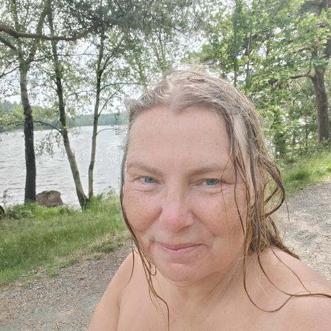 Hed and shoulders of a wet white middle aged woman. Behind her are trees and bushes, and then the lake.