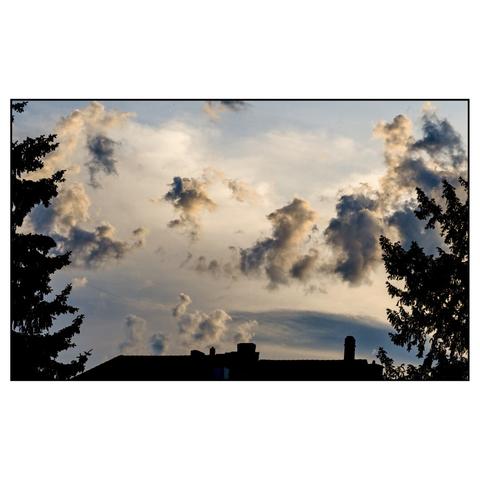 a dramatic evening sky with various clouds forming complex textures, framed by silhouettes of trees on the left an right, and by a roof top on the bottom.
