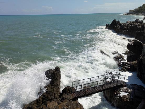 un tratto di costa rocciosa a Cefalù, con le onde del mare che si infrangono con forza contro gli scogli. L'acqua è di un verde-azzurro intenso, tipico del Mar Tirreno, e si estende fino all'orizzonte.

In primo piano si nota una passerella in metallo che collega due formazioni rocciose. La struttura sembra robusta, ma è circondata da mare agitato e schiuma bianca, per via del mare piuttosto mosso