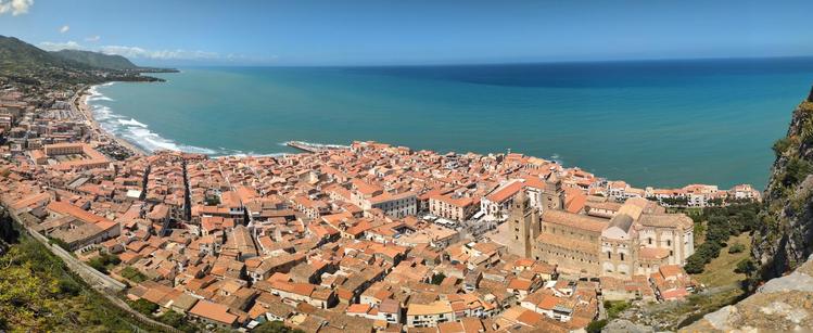 In questa foto vediamo il borgo siciliano affacciato sul Mar Tirreno, fotografato dall’alto, dal promontorio della Rocca di Cefalù. Le casette con i tetti rossi si accalcano in un dedalo di viuzze strette, che si aprono verso il lungomare e il porto.

Il mare è calmo e di un azzurro intenso, che sfuma all’orizzonte in tonalità più profonde. Lungo la costa, si può notare la spiaggia sabbiosa che si estende verso il lato sinistro della foto, dove si intravedono anche le zone residenziali più moderne.