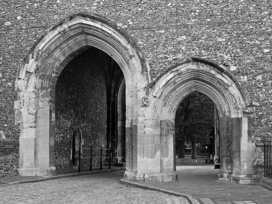 Black and white photo of a passageway framed by two pointed stone arches which share a central pier. The arch on the left is the larger of the two and spans a narrow street. A sidewalk passes beneath the arch on the right. The masonry structure surrounding the pair is composed of flint cobbles interspersed with a smaller quantity of Roman bricks.