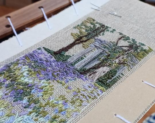 Part worked stitched artwork depicting woodland with bluebells in the foreground and a pillared, stone folly amid the trees. Viewed on an angle to show texture