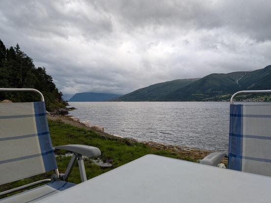 The image depicts a serene lakeside scene with a cloudy sky overhead. In the foreground, there are two folding chairs with blue and white striped fabric, positioned on a grassy area near the water's edge. A white table is placed in front of the chairs, suggesting a spot for relaxation or dining. The calm water reflects the overcast sky, and the mountains in the background are partially obscured by clouds, adding to the tranquil atmosphere. The landscape is lush with greenery, and a small settlement is visible in the distance, nestled among the hills. The overall scene conveys a sense of peacefulness and natural beauty.