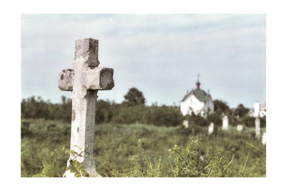 An old stone cross stands in a rural cemetery, surrounded by wild greenery, with a distant, softly blurred church on the horizon. Shot on expired Kodak Gold 100 from 1995, evoking a sense of transience and memory lost to time.