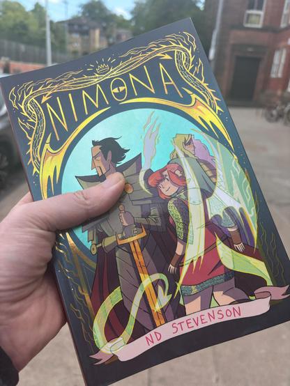 Nimona 10th Anniversary limited edition cover, with shiny gold lettering. Lord Ballister Blackheart looks contemplative, while Nimona smirks at the camera