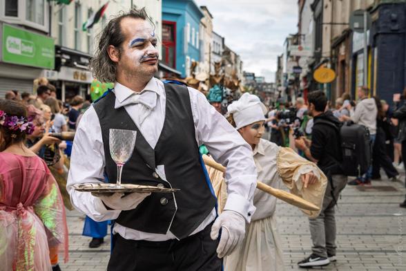 A lively street parade scene in Cork during The Family Shenanigans parade, featuring a man in theatrical waiter costume with white gloves, face paint, and a bow tie, holding a silver tray with a wine glass, surrounded by other performers in colourful costumes and a bustling crowd in the background.