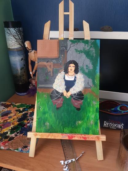 Photo d'une peinture à l'acrylique sur un chevalet. Est représenté une personne, la peau blanche, les cheveux noirs et bouclés, les yeux bleus et du far à paupière et rouge à lèvres bordeaux. Iel a plusieurs piercing, un au menton, un au dessus de son sourcil gauche. 
Iel est accroupie dans l'herbe fleurie devant un atelier de bricolage dans une maison en ciment grise.
Iel est habillé d'un t-shirt blanc, du haut d'une salopette, d'un pantalon noir et bordeau, de chaussures noires et du veste très froufrous multicolores. Iel a deux chaines fines autour du cou, et plusieurs qui décore son pantalon.