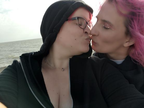 Zwei Personen sind auf dem Bild zu sehen. Links Ari mit roten Haaren und schwarzer Kapuzenjacke, rechts Danielle mit pinken Haaren ..beide haben die Augen zu und küssen sich. Im Hintergrund sieht man die Nordsee