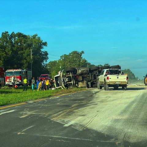 Overturned rock-hauling truck blocking the left lane and median of a highway with spilled rock on the highway.