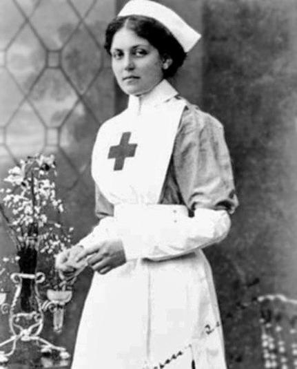 Eine junge Frau in einer historischen Krankenschwester-Uniform mit einem roten Kreuz auf der Brust. Sie hat einen weißen Haarschleier und steht aufrecht, während sie in die Kamera blickt. Im Hintergrund sind Fenster mit dekorativem Muster sowie ein Tisch mit einer Vase und Blumen zu sehen.